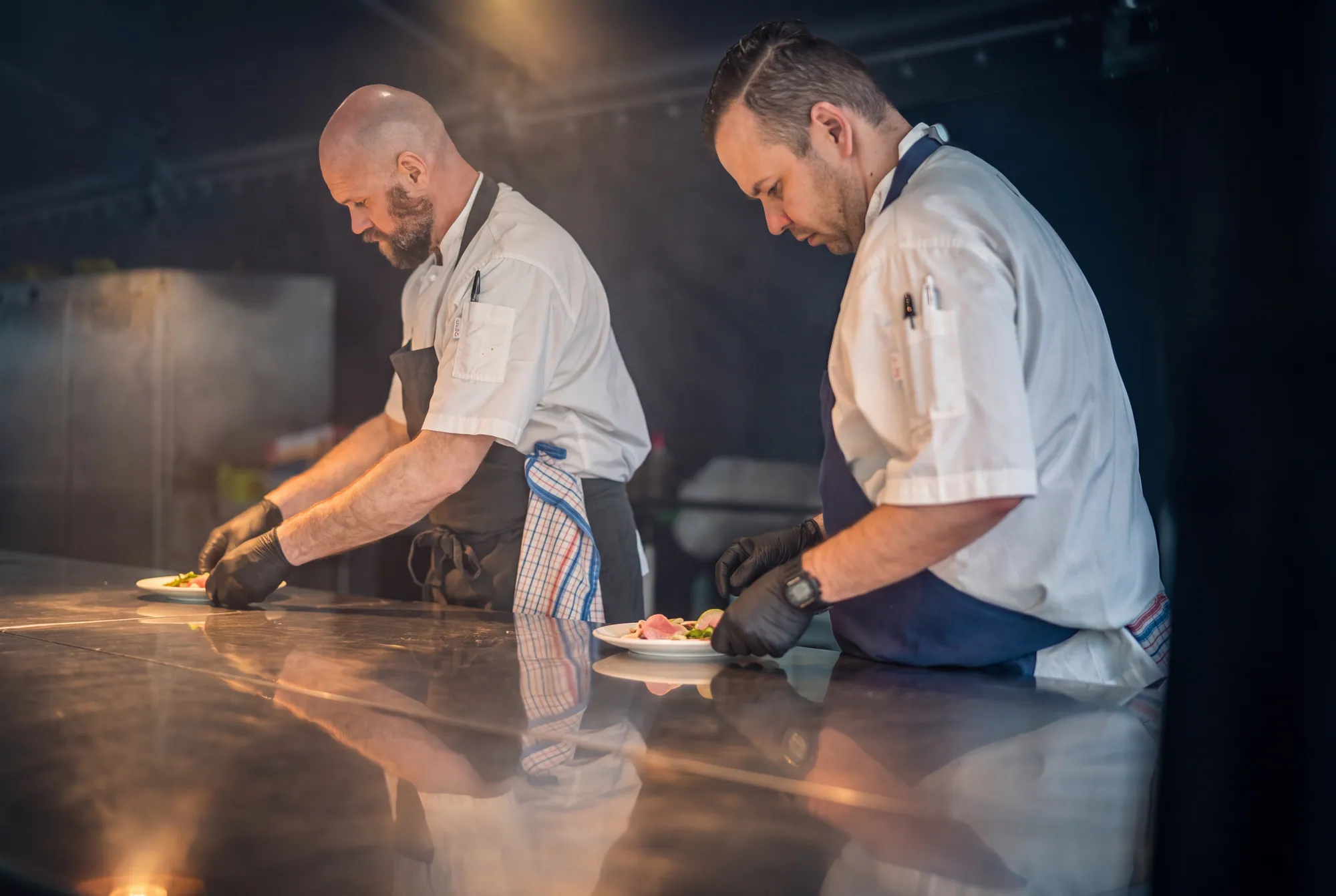 Two chefs plating at a steel bench