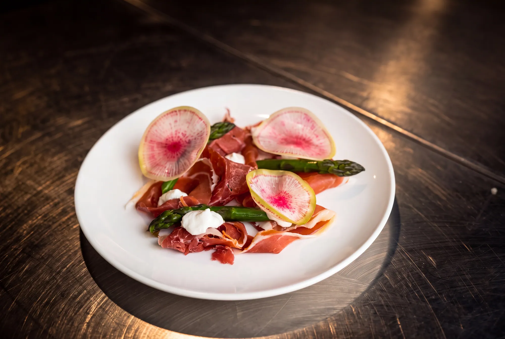 Prosciutto, asparagus and watermelon radish entrée on a steel bench